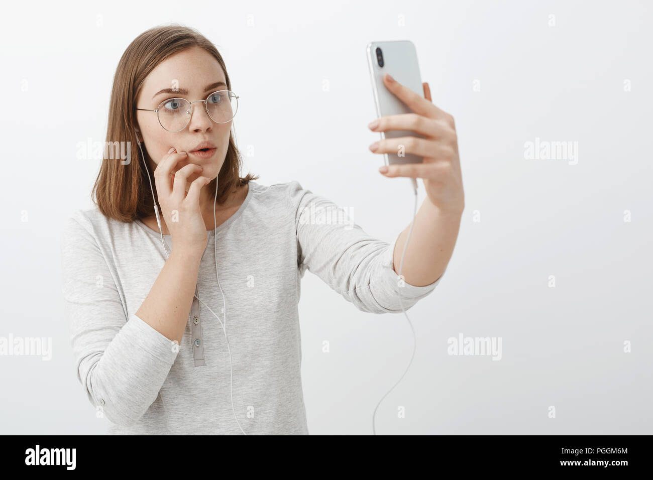 Waist-up shot of creative curious and entertained charming woman in glasses wearing earphones raising hand with smartphone gazing intrigued and interested at device screen watching amazing video Stock Photo