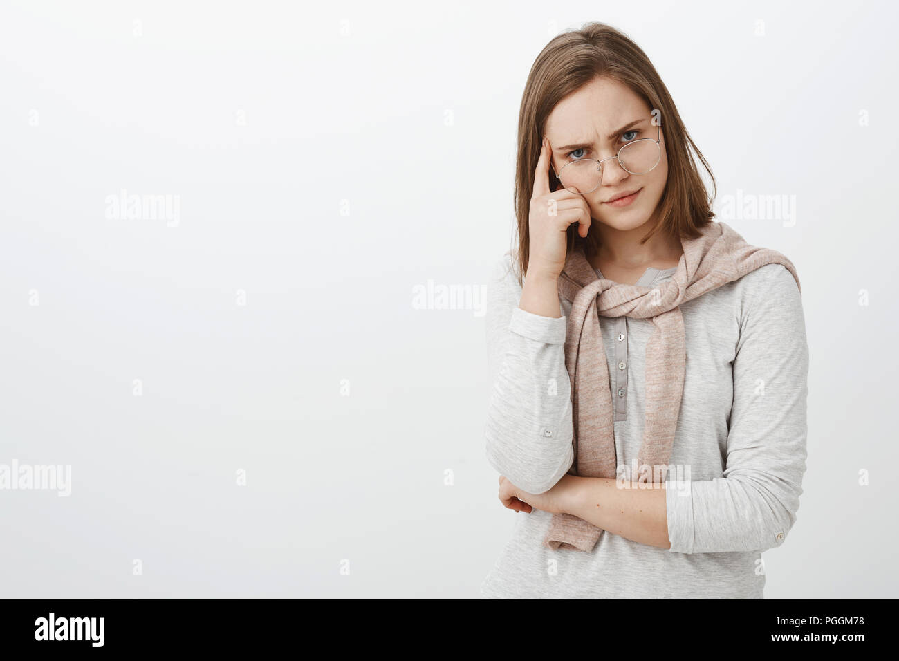 Displeased intense and tired female tutor in glasses frowning looking from under frames holding finger on temple thinking person is dumb losing temper from irritation and annoyance over grey wall Stock Photo