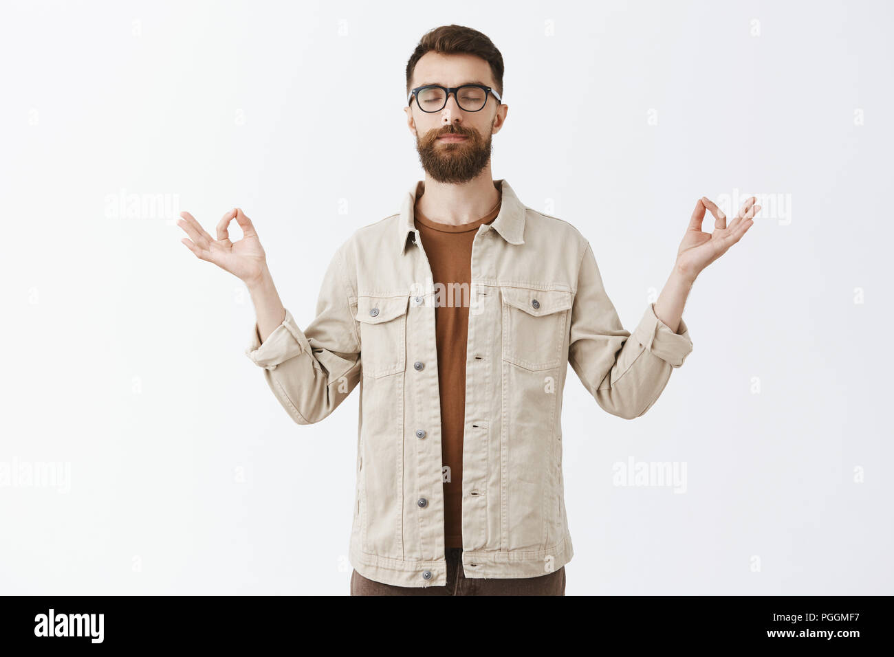 Calming down with yoga. Portrait of relieved peaceful good-looking urban guy in jacket with long sick beard closing eyes spreading hands in zen gesture meditating to restore peace and patience Stock Photo
