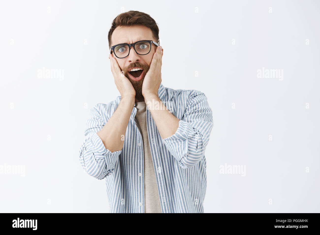 Waist-up shot of impressed and surprised sociable good-looking male with long beard and moustache holding palms on cheeks and staring at camera hearing shocking rumor over gray background Stock Photo