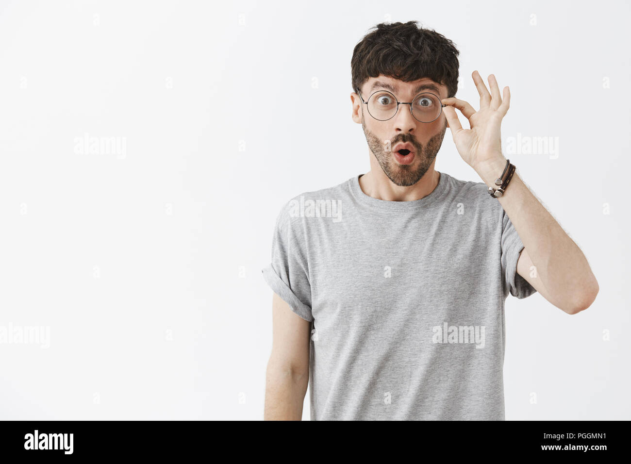 Waist-up shot of enthusiastic impressed dark-haired guy with beard in stylish transparent glasses touching rim of eyewear and folding lips from joy and excitement staring at amazing advertisement Stock Photo