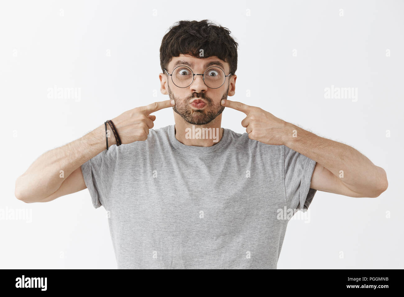 Man never losing sense of humour and optimism. Portrait of happy childish and attractive young father with beard in glasses pouting and poking blowed cheeks smiling, having fun over gray wall Stock Photo