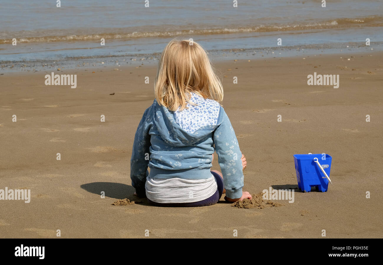 Small girl sitting on sand beach at low tide with blue bucket. Stock Photo