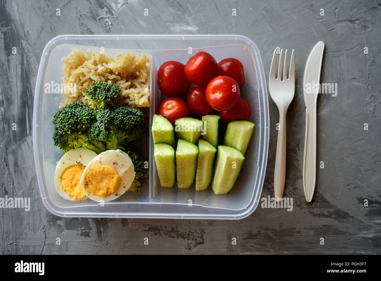 Lunch box with healthy food. Rice, broccoli, tomato, cucumber, eggs, apple and water. On grey concrete background Stock Photo