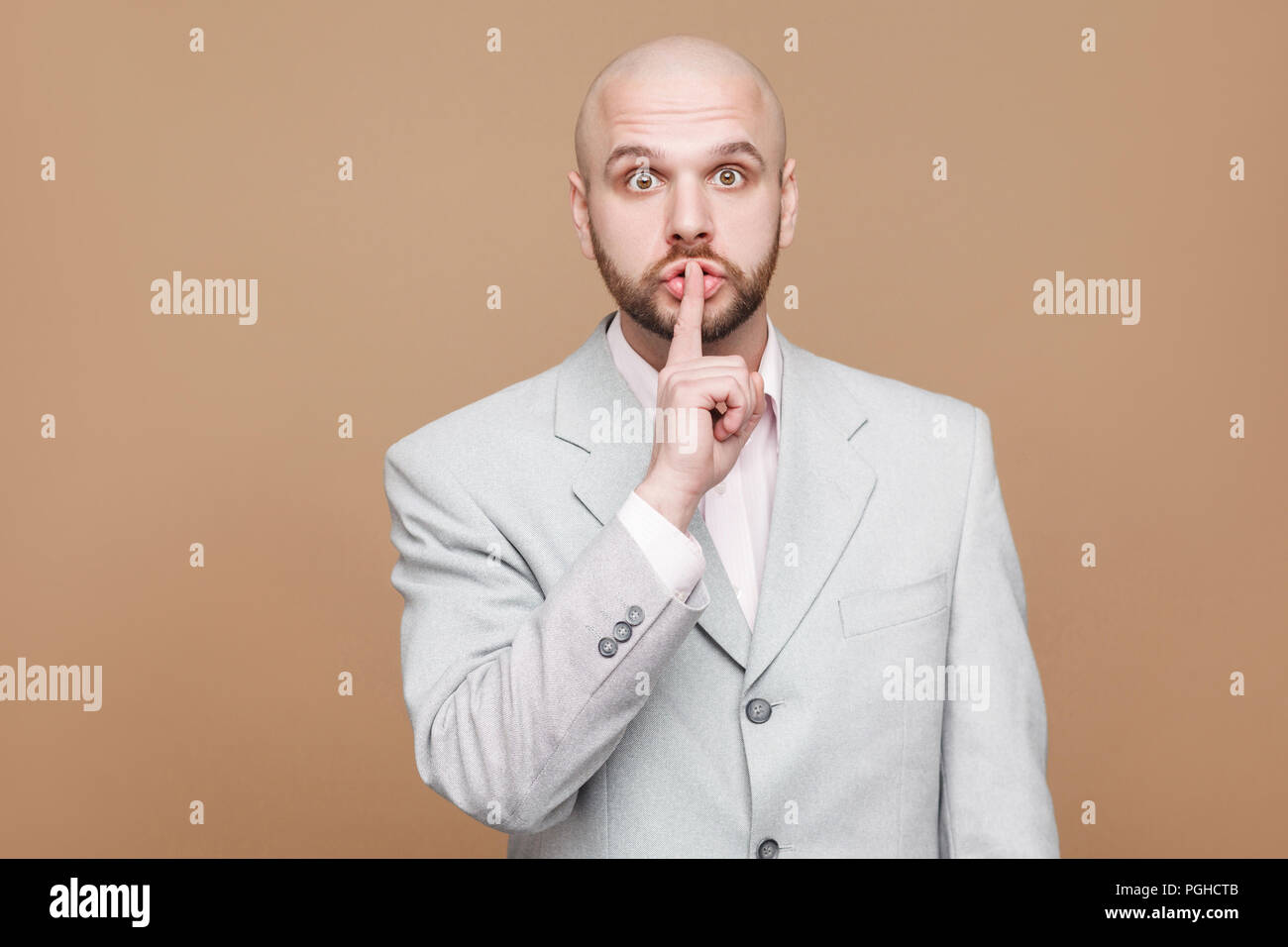 shhh this is secret. Portrait of handsome middle aged bald bearded businessman in classic light gray suit standing and looking at camera with silence  Stock Photo