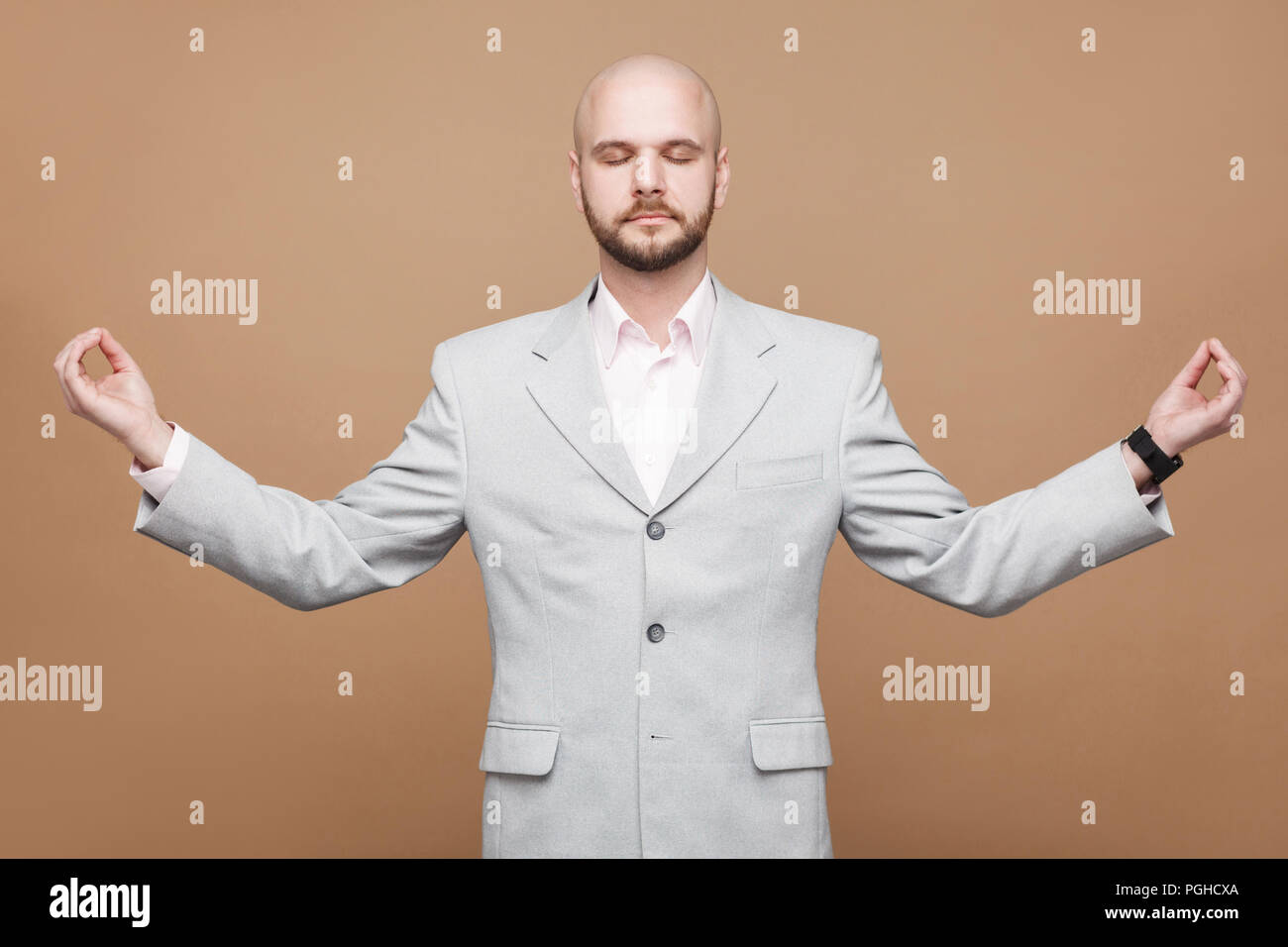 Portrait of calm handsome middle aged bald bearded businessman in classic light gray suit standing with yoga pose raised arms and clossed eyes. indoor Stock Photo