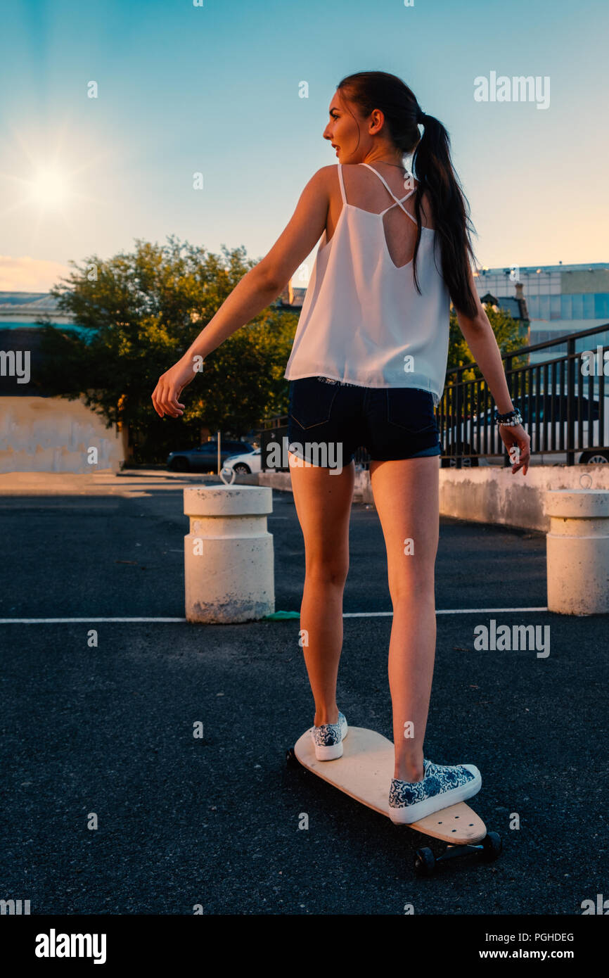 Beautiful girl riding skateboard in short denim shorts, view from back