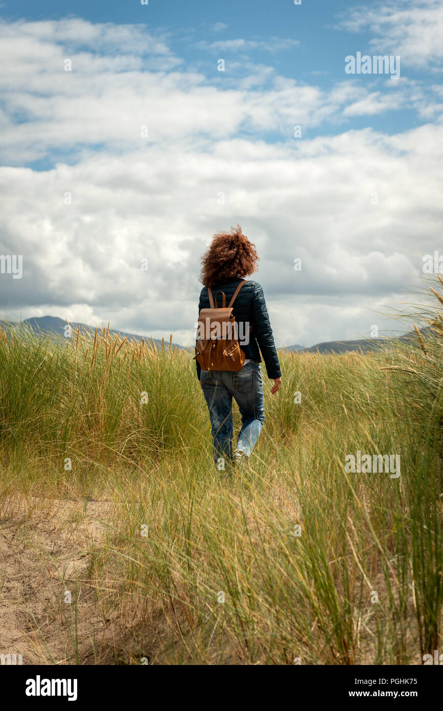 Young beautiful woman with red curly hair wearing casual  blue jacket and blue jeans walking on coastal sand dune through marram grass on a sunny day. Stock Photo
