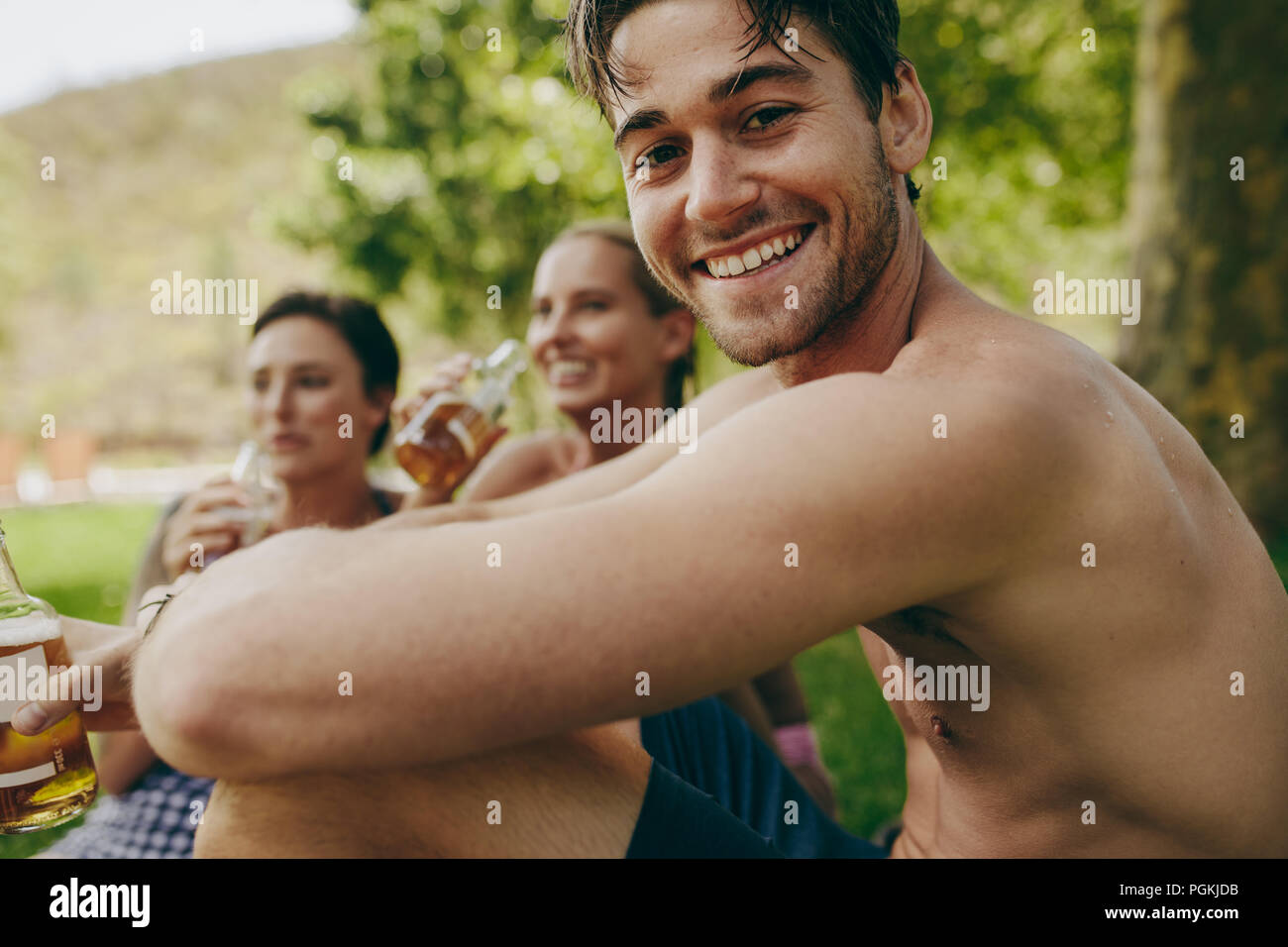 Friends enjoying holiday at a park with drinks. Man with two female friends sitting outdoors drinking beer. Stock Photo