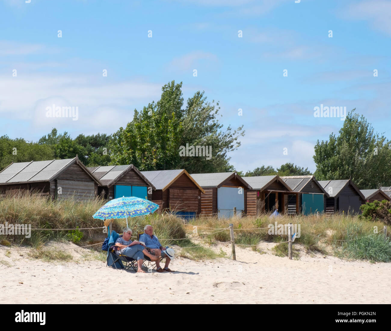 A couple sitting under an umbrella next to beach huts on the beach at Studland Bay, Jurassic Coast, Dorset, UK Stock Photo