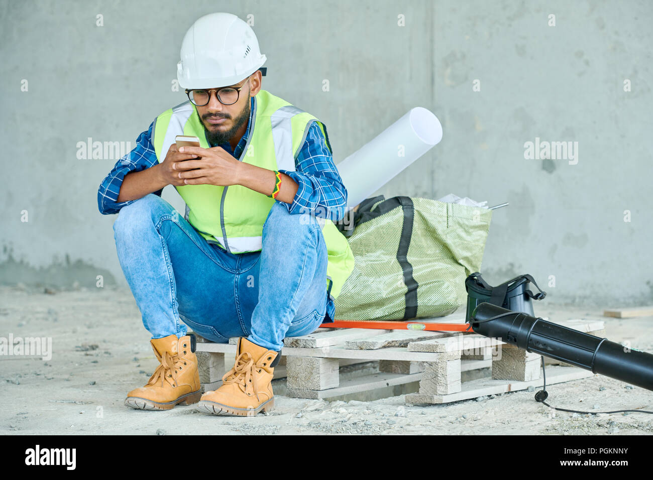 Full length portrait of Middle-Eastern construction worker using smartphone while taking break from work on site, copy space Stock Photo