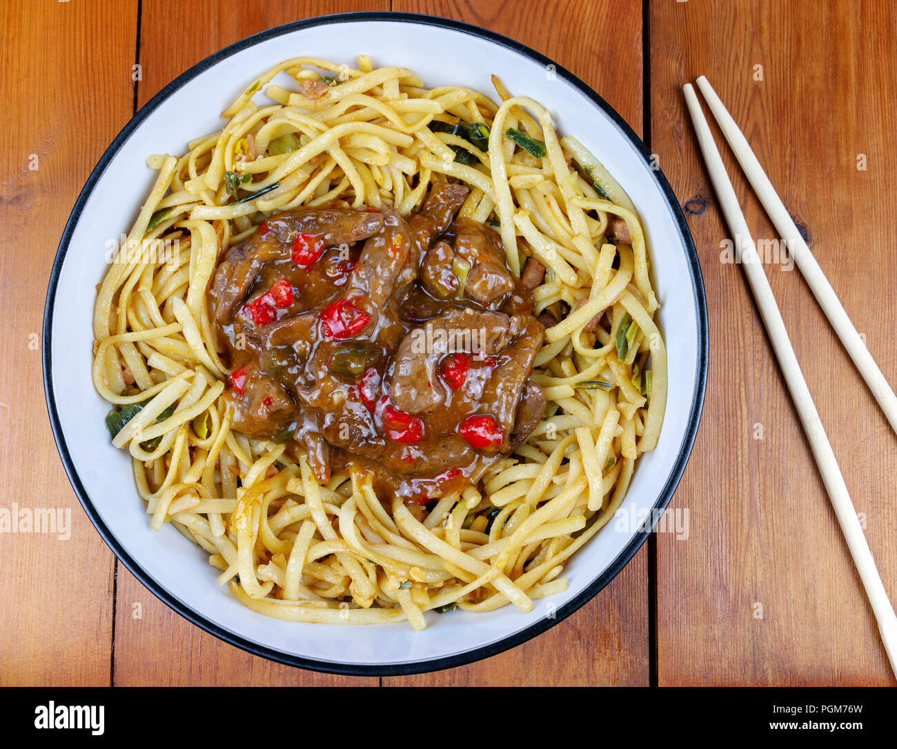 Spicy chilli beef with fried noodles in a dish with chopsticks on a wooden table from an overhead perspective Stock Photo