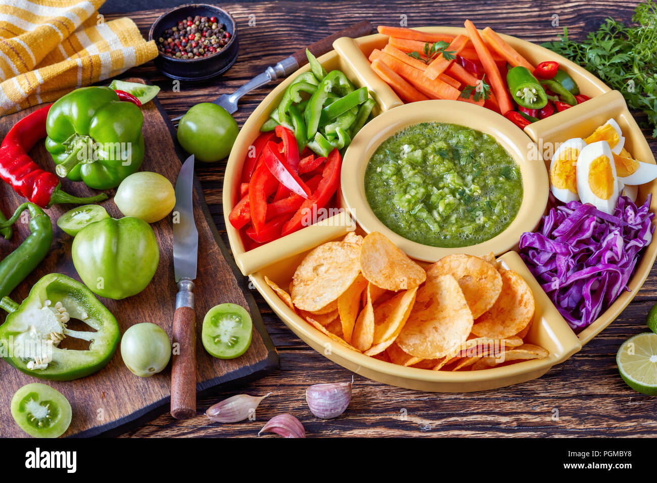 green salsa verde and mexican set of chopped fresh vegetables, hard boiled eggs and chips in bowls on old rustic wooden board with ingredients, view f Stock Photo