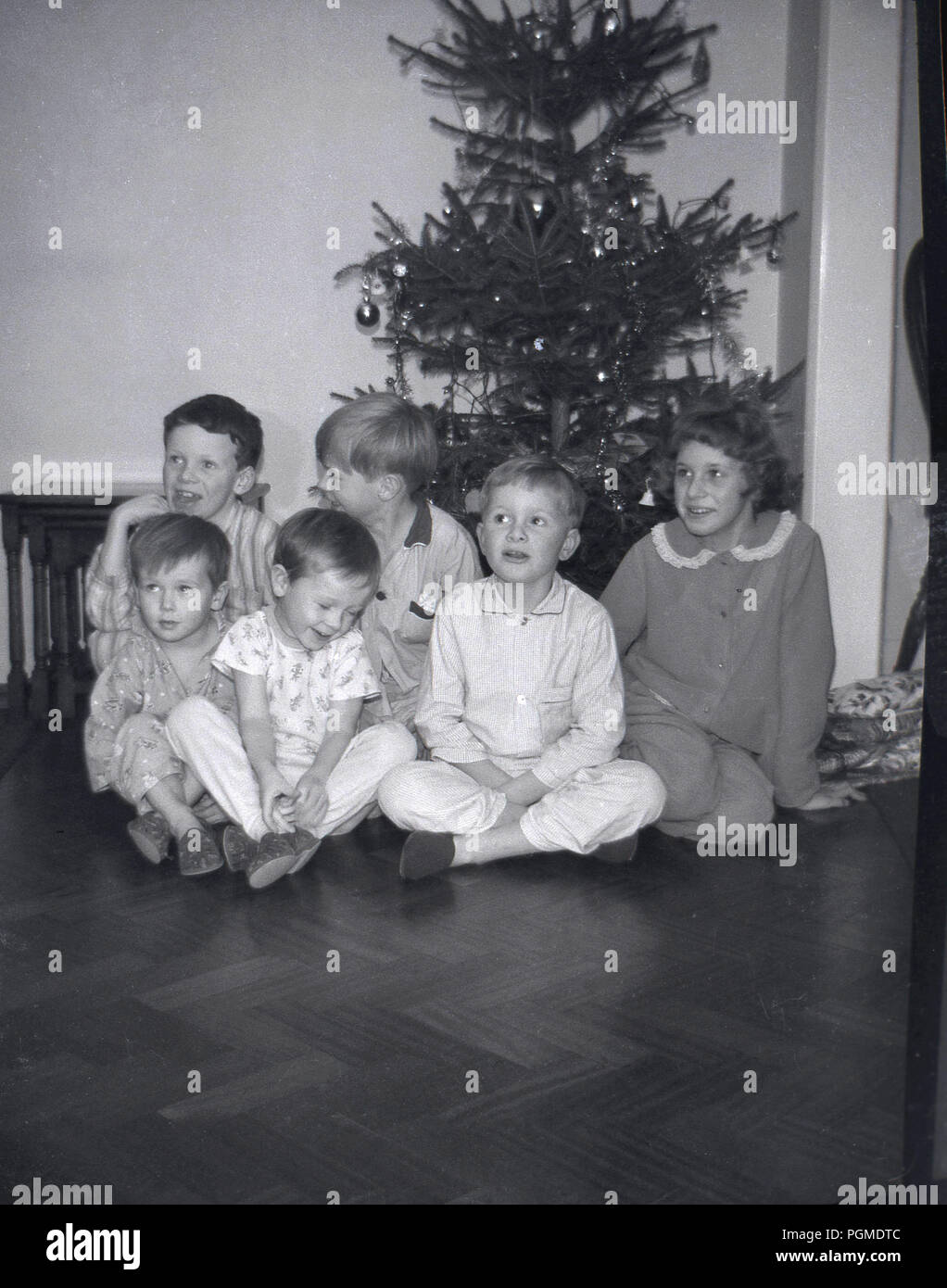 1960s, histroical, Xmas morning and a group of young children in their pyjamas and slippers sit together inside on a wooden floor infront of a christmas tree, England, UK. Stock Photo