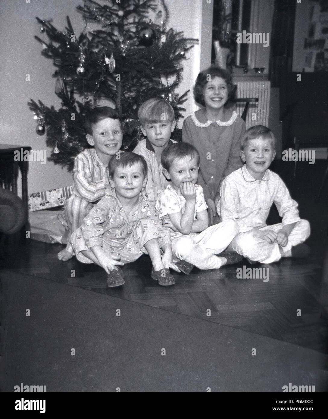 1960s, histroical, Xmas morning and a group of smiling young children in their pyjamas sit together inside on a wooden floor infront of a christmas tree, England, UK. Stock Photo