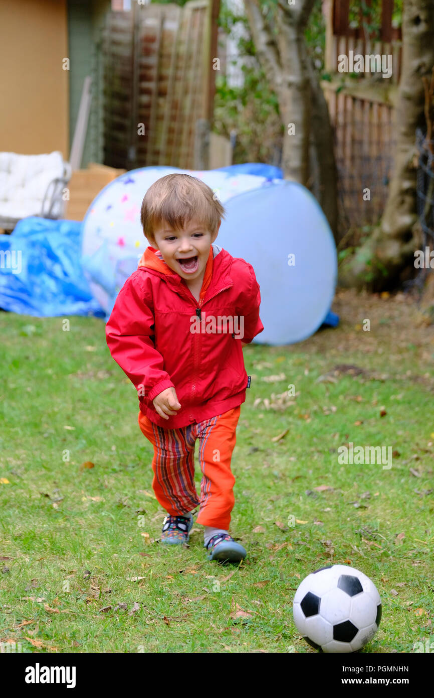 Boy aged two (2) with red coat and football happy and smiling as he plays with football in garden Stock Photo