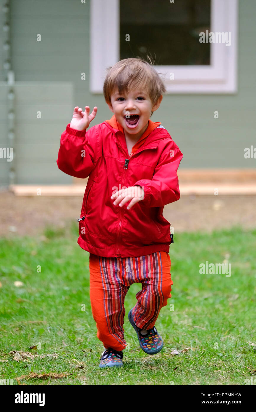 Boy aged two (2) with ecited expression on face as he runs in red coat outside in garden Stock Photo