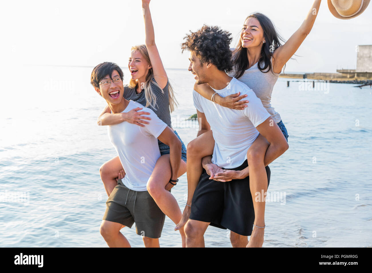Group of friends walking along the beach, with men giving piggyback ride to girlfriends. Happy young friends enjoying a day at beach Stock Photo