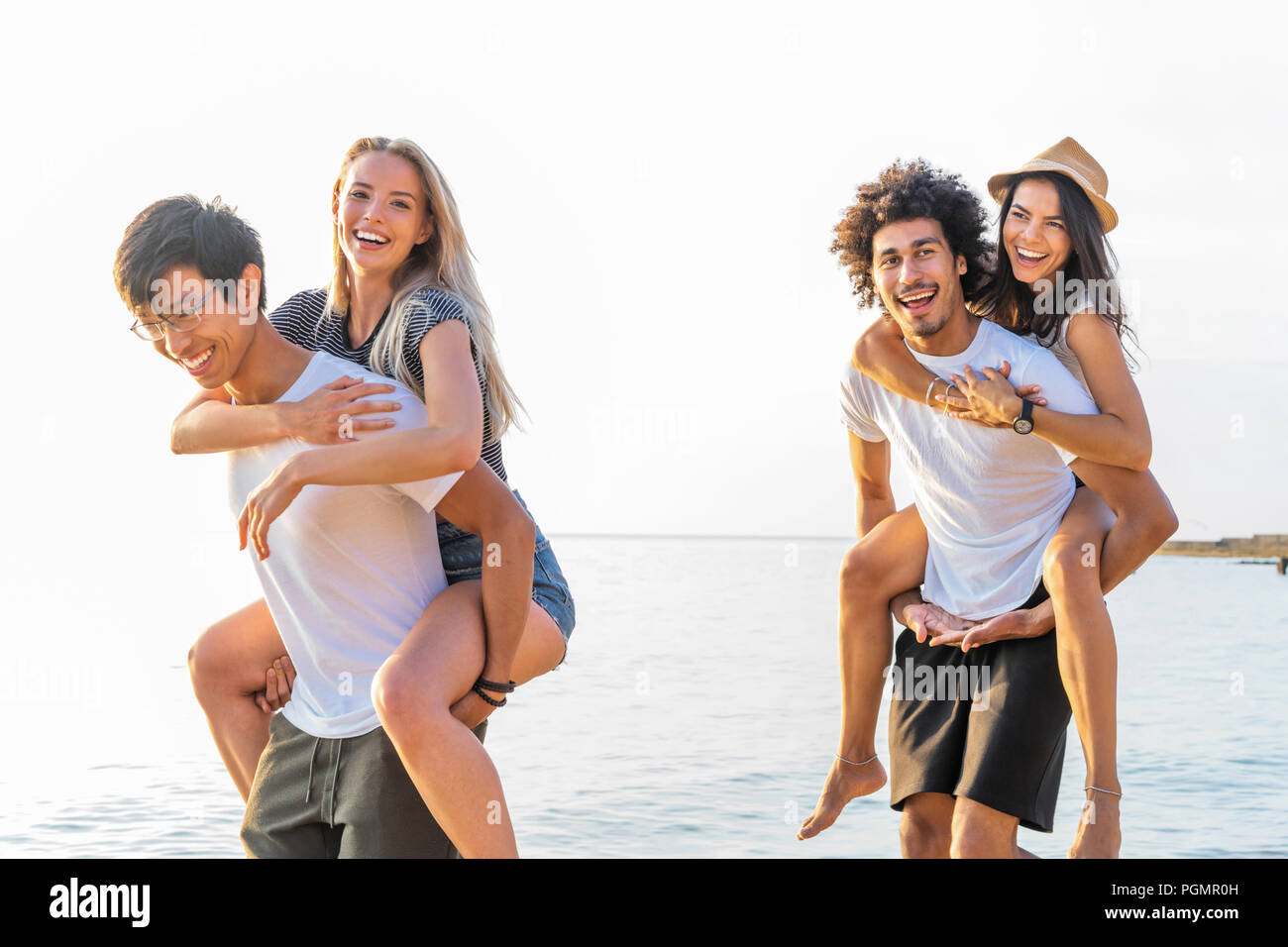 Group of friends walking along the beach, with men giving piggyback ride to girlfriends. Happy young friends enjoying a day at beach Stock Photo