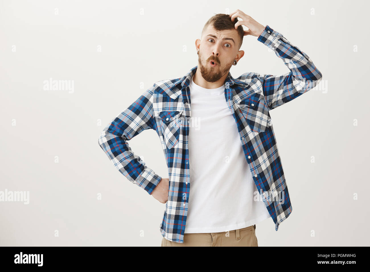 Funny good-looking caucasian male student with beard in blue plaid shirt, preparing for exam, scratching head, looking confused and clueless, having no idea what to answer, questioned over gray wall Stock Photo