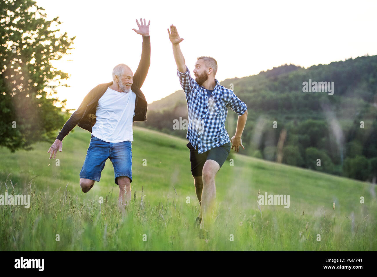 An adult hipster son with his senior father jumping on a meadow in ...