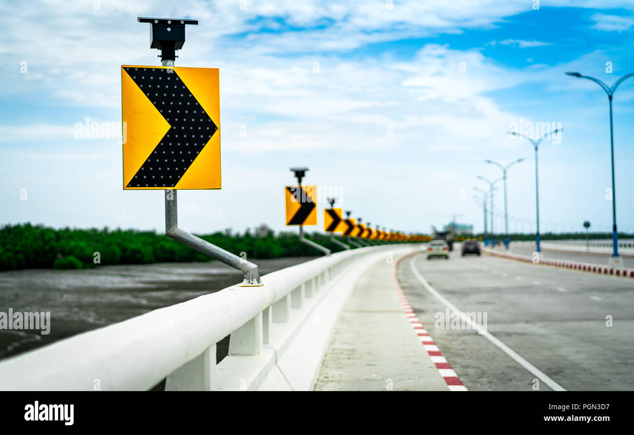 Black and yellow arrow on curve traffic sign on the bridge with solar cell panel ob blurred background of concrete road and car near mud flat and mang Stock Photo