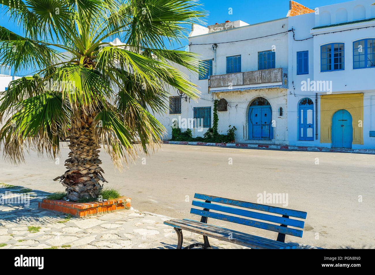 The seaside promenade with lush palms and small benches and a view on the old houses, stretching along the coast, Mahdia, Tunisia. Stock Photo