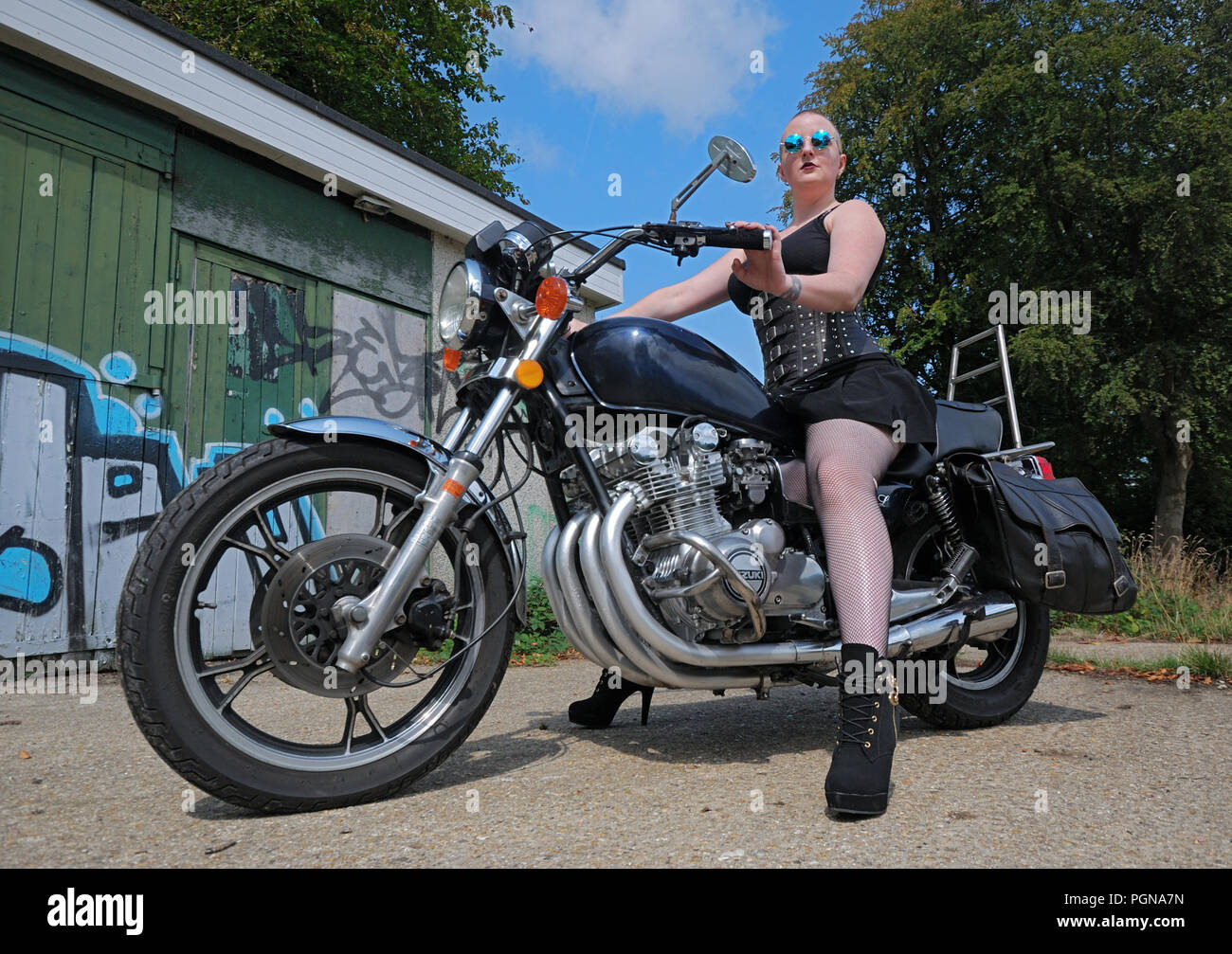 Model Rosa Fury straddles a classic Suzuki motorcycle in front of a graffiti covered wall. She's wearing a short black dress with a corset & fishnets. Stock Photo