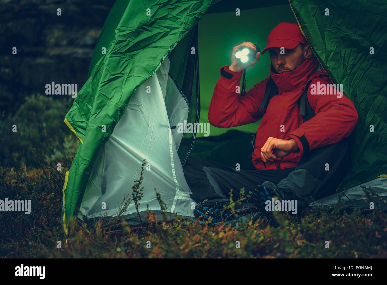 Caucasian Camper with Flashlight Inside His Tent During Rainy Evening While on Hike. Stock Photo