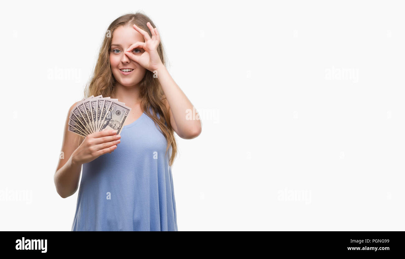Young blonde woman holding dollars with happy face smiling doing ok sign with hand on eye looking through fingers Stock Photo