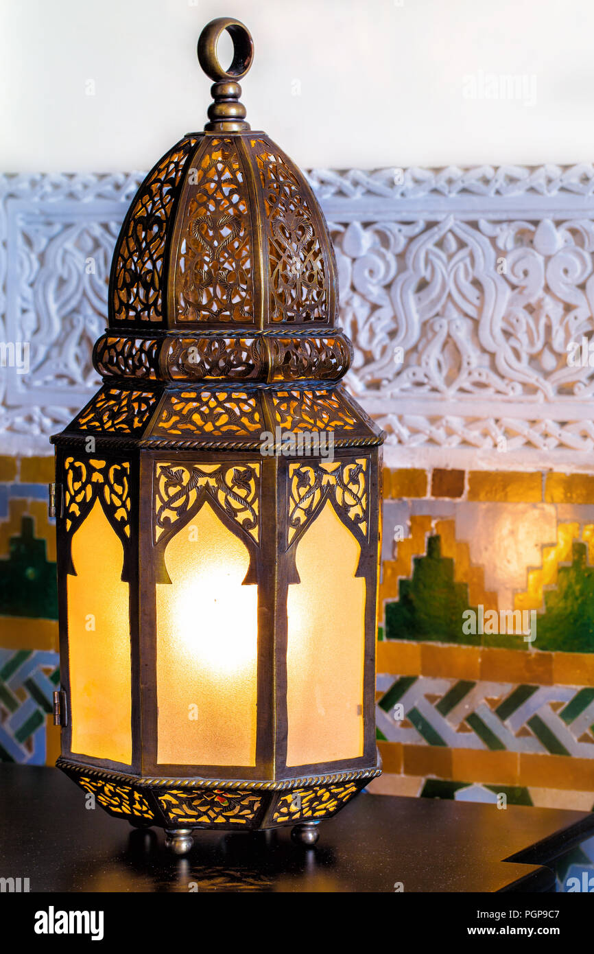 Moroccan metal lantern converted into a wired small table lamp. Shown illuminated, with a soft glow lighting the mosaic tile wall behind it. Marrakech. Stock Photo