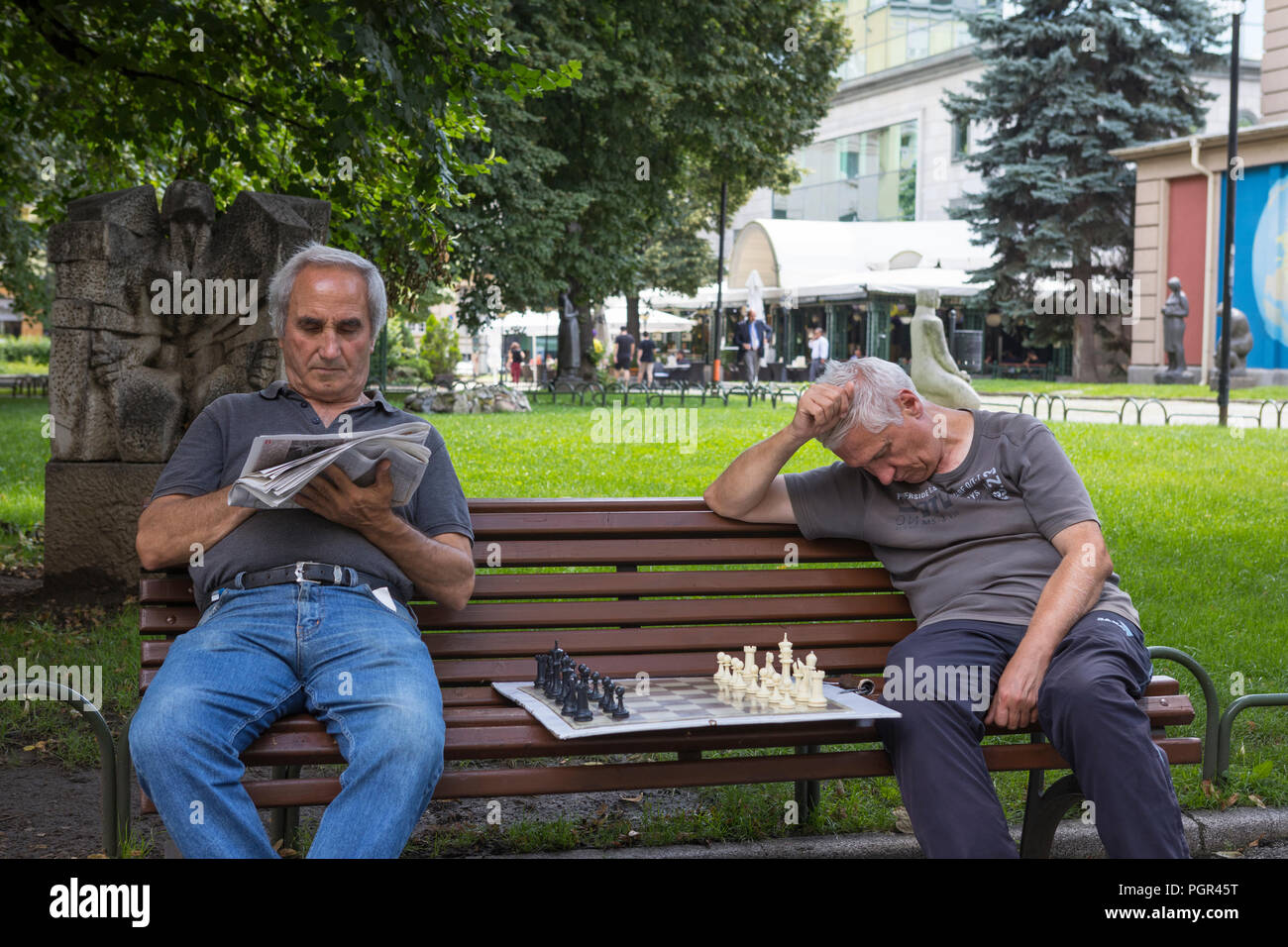Two men resting at a bench, one reading, one fallen asleep with a chessboard in the middle, Sofia, Bulgaria Stock Photo