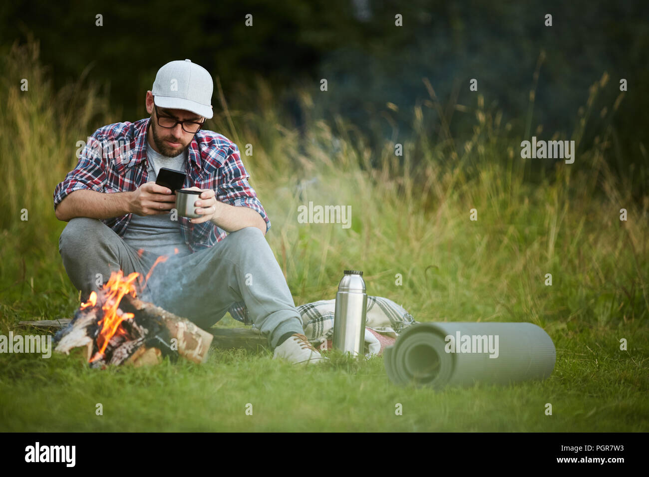 Young man drinking tea and using his smartphone by campfire Stock Photo