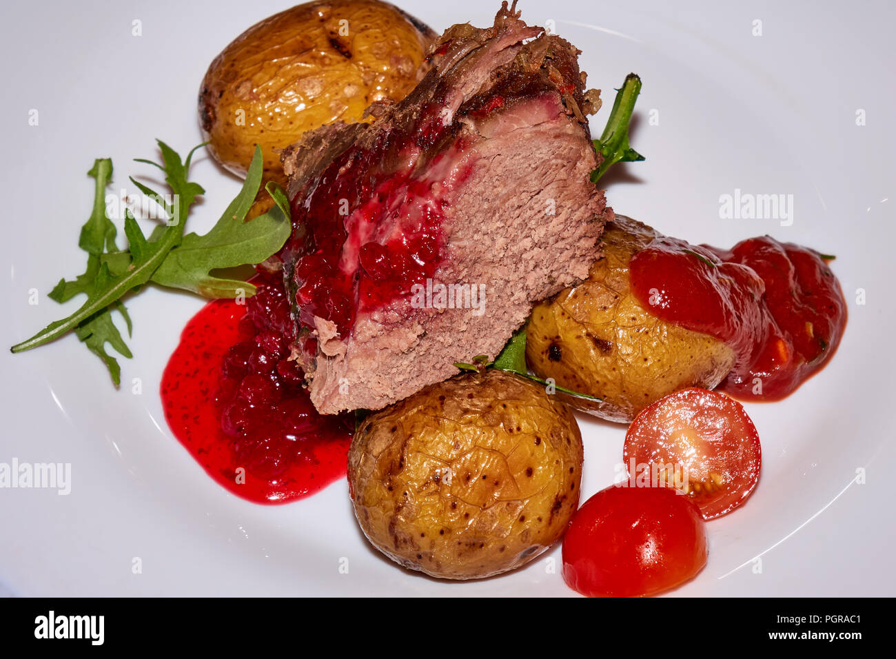 Big peace of the roasted medium well mutton meat with baked potatoes, fresh tomatoes and cowberry sauce on the white plate Stock Photo