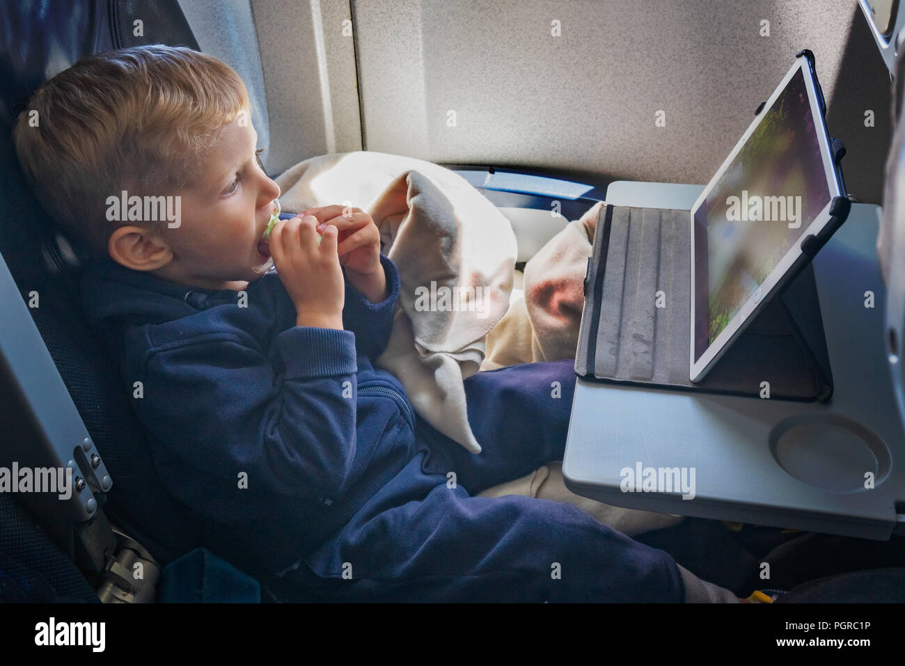 Baby boy is sitting on the plane and eats and looks at the tablet in front of him. Stock Photo