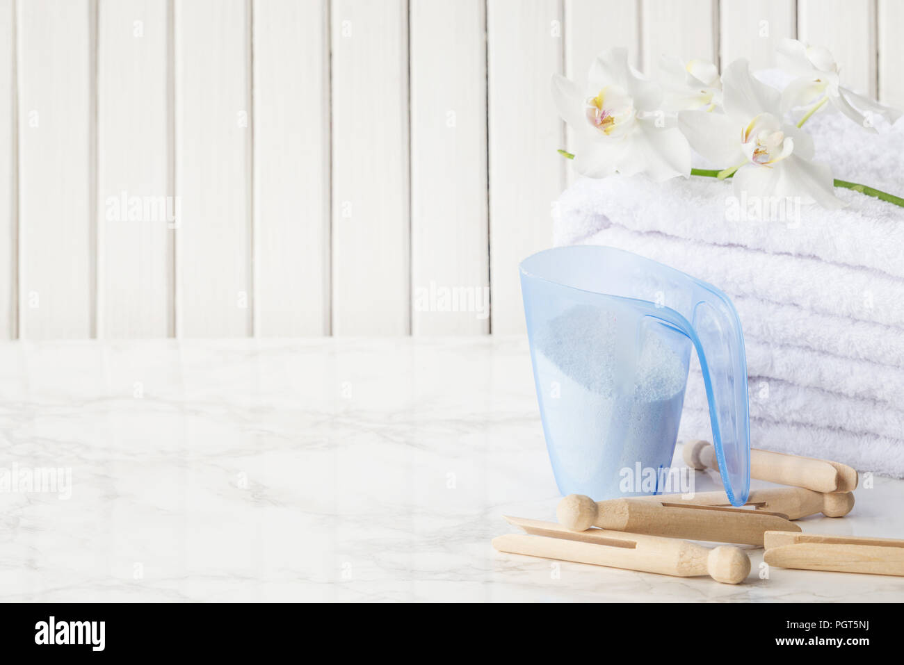 Blue plastic measuring beaker with detergent, stack of white terry towels, wooden clothespins and white orchid flower are on a marble surface against  Stock Photo