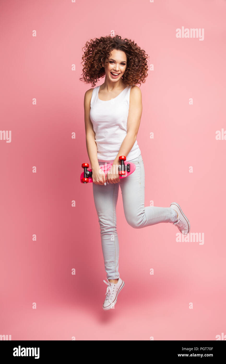 Full length portrait of a beautiful happy woman in summer clothes posing and looking away while jumping and holding skateboard isolated over pink background. Stock Photo