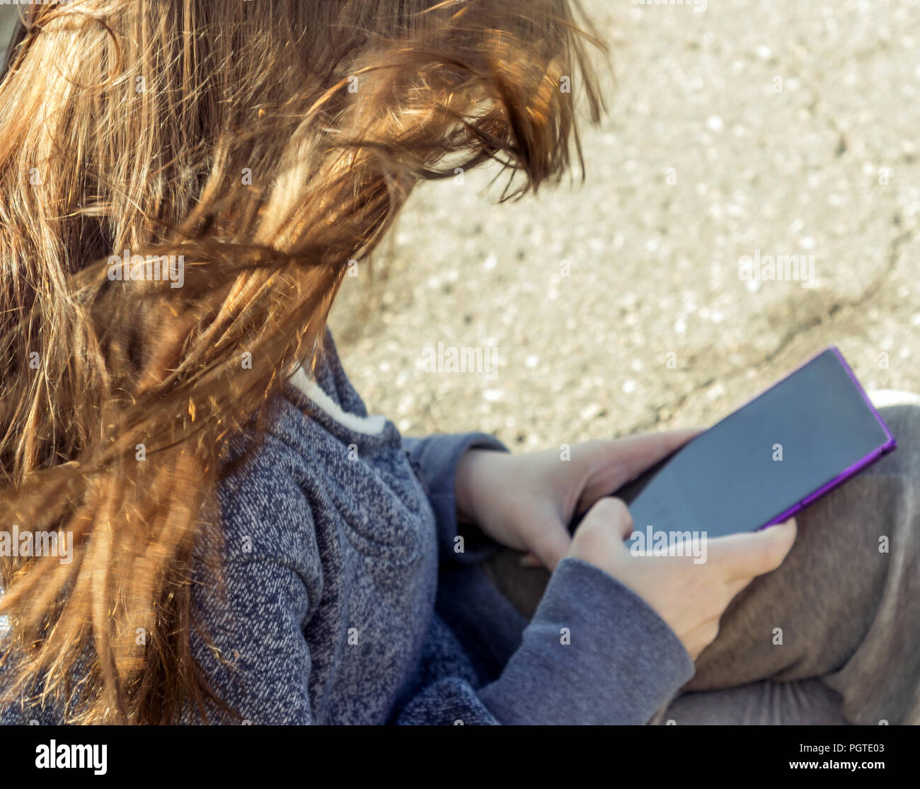 the girl is sitting with a large phone in the hands of a lilac color, her hair is cinnamon-red is developing in the wind, a blue jacket, a sunny day, Stock Photo
