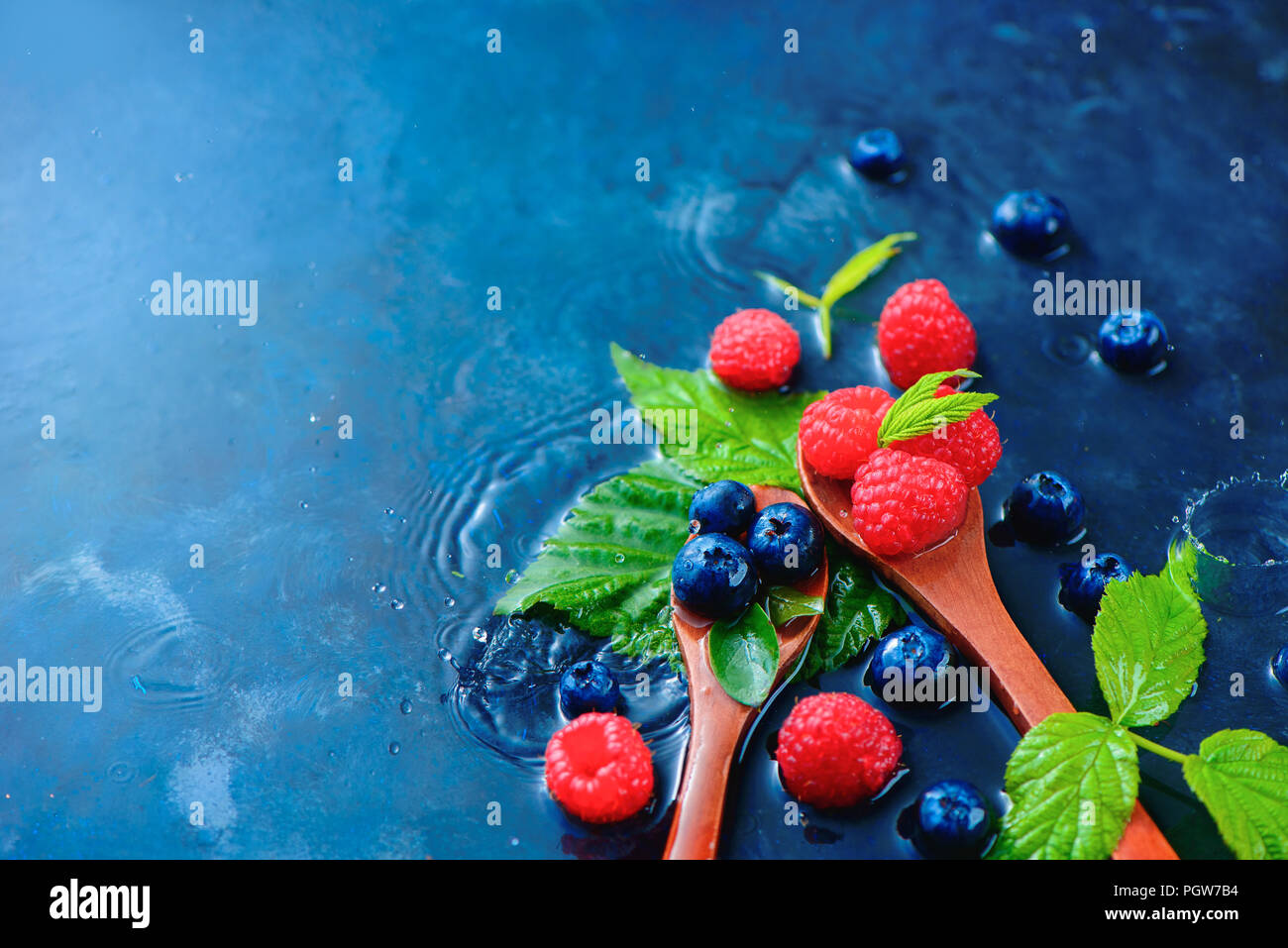 Summer berries in wooden spoons with raindrops. Raspberry and blueberry mix on a dark blue wet background with copy space. Raw ingredients concept Stock Photo