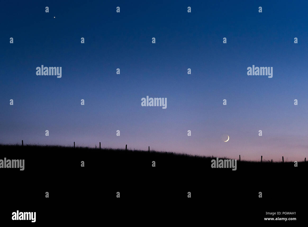 The skyline cradles the crescent moon in the pale glow of the pastel twilight below Venus, an old fence line is visible along the gentle rolling hills. Stock Photo