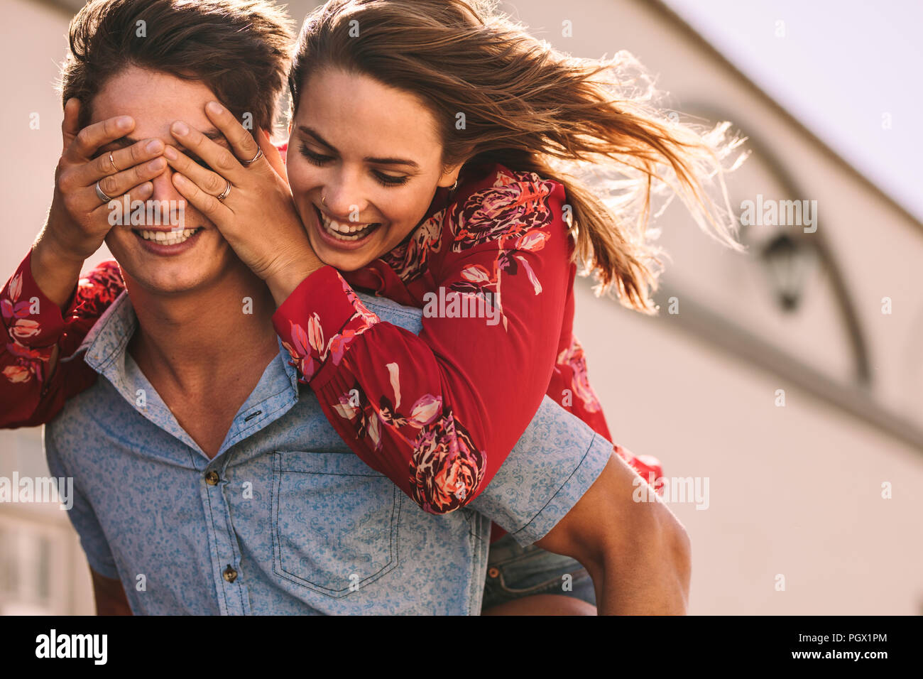 Smiling woman piggy riding on man closing his eyes with her hands. Smiling man carrying woman on his back walking outdoors. Stock Photo