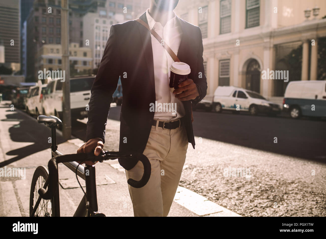Cropped shot of young businessman going to work by bike. Man in suit holding a cup of coffee walking by the street with his bicycle. Stock Photo
