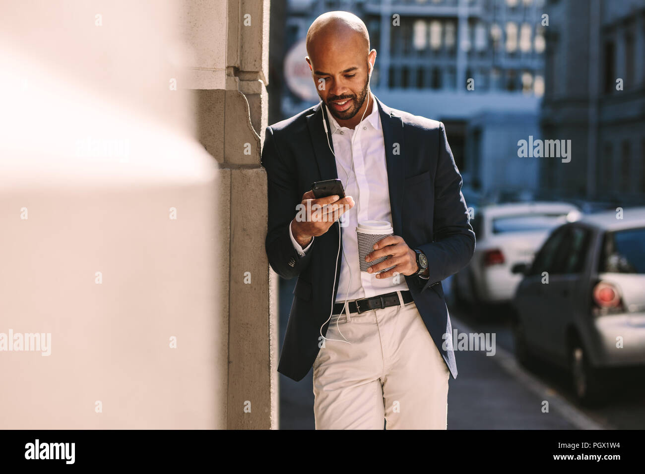 Handsome businessman leaning on a wall while standing outdoors and using smartphone. Man in suit wearing earphones looking at his mobile phone. Stock Photo