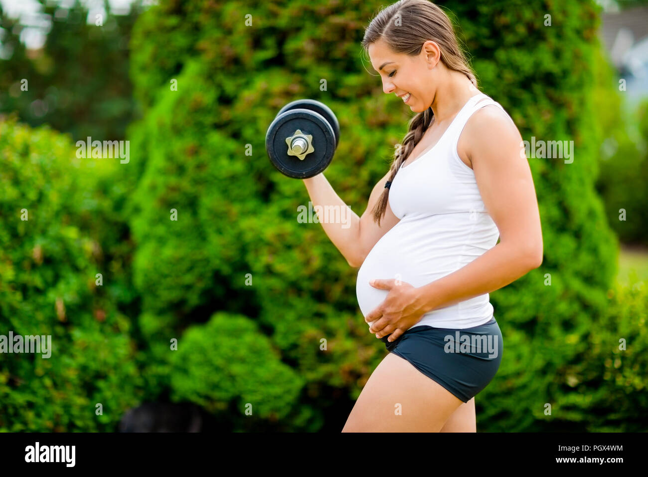 Expectant Mother Touching Belly While Lifting Dumbbell In Park Stock Photo