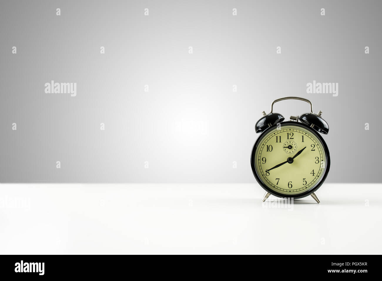 Vintage style metal alarm clock with bells standing on a white counter against a graduated grey background with central highlight and plenty of copysp Stock Photo