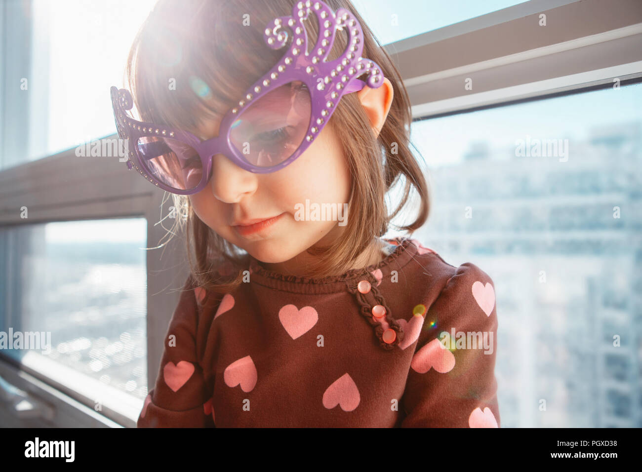 Portrait of Caucasian girl child sitting on window sill at home and blowing whistle trumpet celebrating birthday. Toddler preschooler wearing funny gl Stock Photo