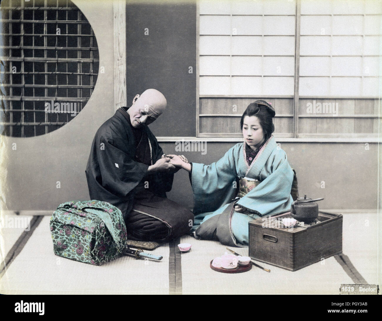 [ 1890s Japan - A Japanese Doctor Takes a Patient’s Pulse ] —   A bald-headed doctor is taking a patient’s pulse. Doctors shaved their heads because medicine were originally administered by Buddhist priests. His young female patient sits next to a hakohibachi, a brazier encased in a wooden box. In front of the box lies what appears to be a kiseru pipe and a tray of tea cups.  19th century vintage albumen photograph. Stock Photo