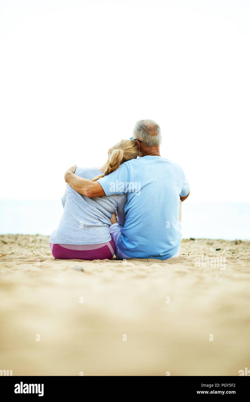 Back view of affectionate senior couple sitting on sand in embrace and relaxing after workout Stock Photo