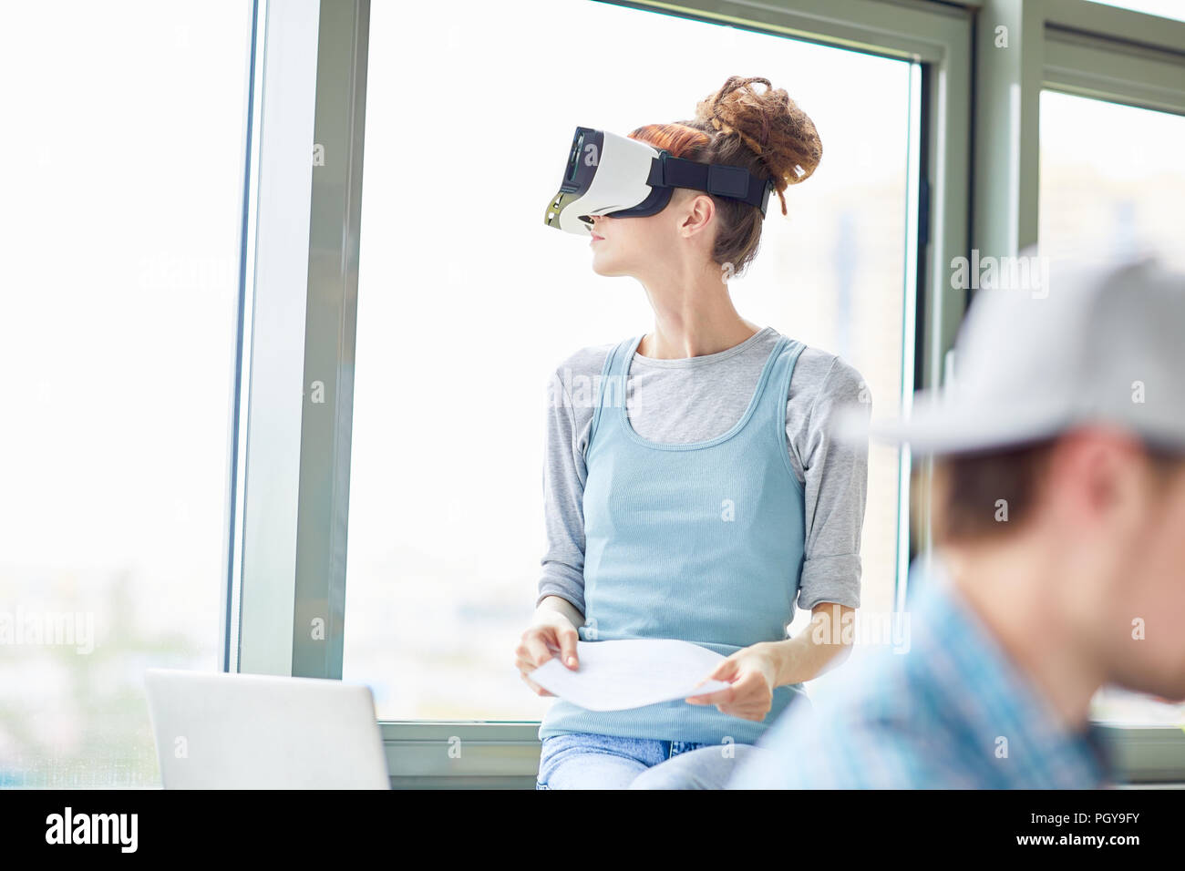Purposeful hipster girl in virtual reality goggles sitting on window-sill and looking out window while thinking about project Stock Photo