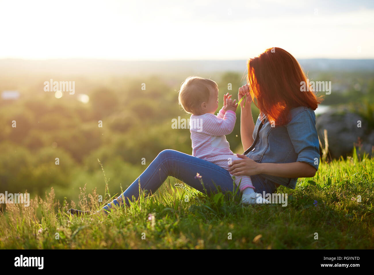 Mother and daughter spending family time sitting on grass while sunset. Mom keeping child on knees, playing, feeling happy, having red hair, wearing b Stock Photo